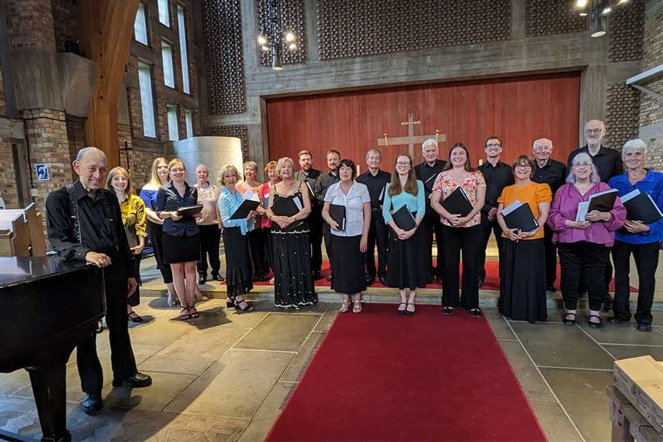 A group of men and women gathered within Keele Chapel, alongside the piano, smiling at the camera.