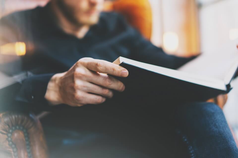 Photograph of a person reading a book with a hand and hard back book in the foreground.