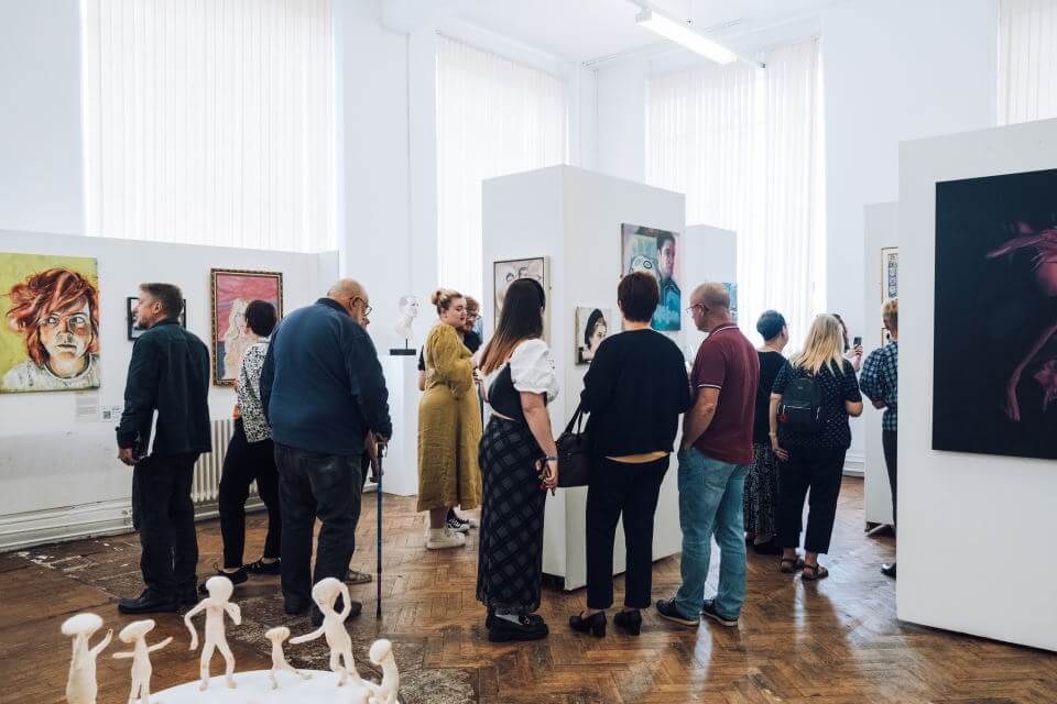 A group of people inside an exhibition looking at artwork on the walls one vivid portrait visible
