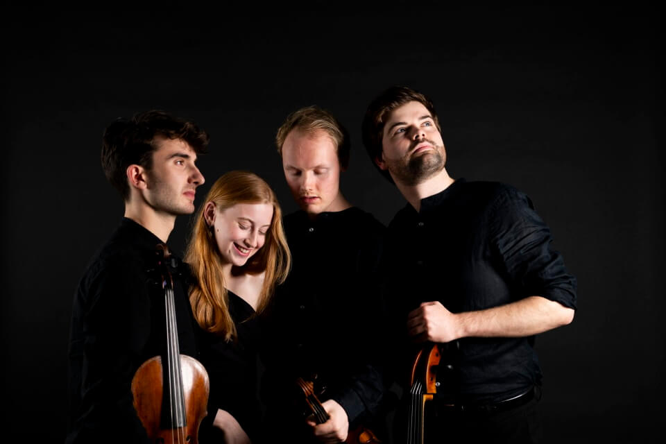 Four performers holding string instruments wearing black on black background 