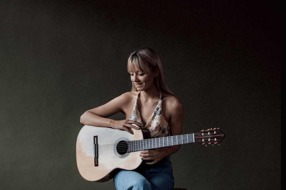 Alexandra holding a guitar seated wearing jeans and halter top smiling and looking down