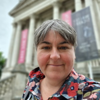 Person looking at you with colourful shirt, smiling. Sort grey hair. Columned building in the background.