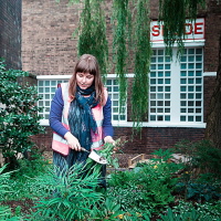 A person tending to a garden, wearing denim and a waistjacket. Long brown hair. With building to the rear brick with white windows.