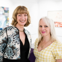 Two women looking forward and smiling. One with chin length brown hair and fringe wearing a mix of black and white other with long white blonde hair wearing yellow and white check..